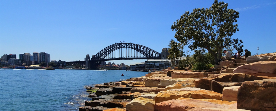 A photograph of Sydney Harbour showing the Sydney Harbour Bridge under a blue sky and sandstone rocks in the foreground.
